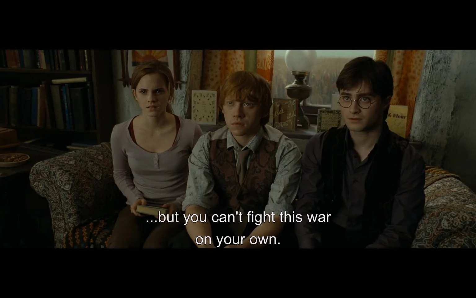 In the scene, three individuals are seated closely together, appearing to be in a tense discussion. The first person, wearing a light purple long-sleeve top, has a concerned expression. The second individual, dressed in a patterned vest over a light shirt, looks worried and attentive. The third person, in a dark vest and glasses, appears serious and focused. The setting is indoors, with a cozy yet cluttered background featuring bookshelves, decorative items, and a lamp. The atmosphere suggests a moment of emotional weight, as indicated by the dialogue displayed on the screen, which emphasizes the importance of not facing challenges alone.