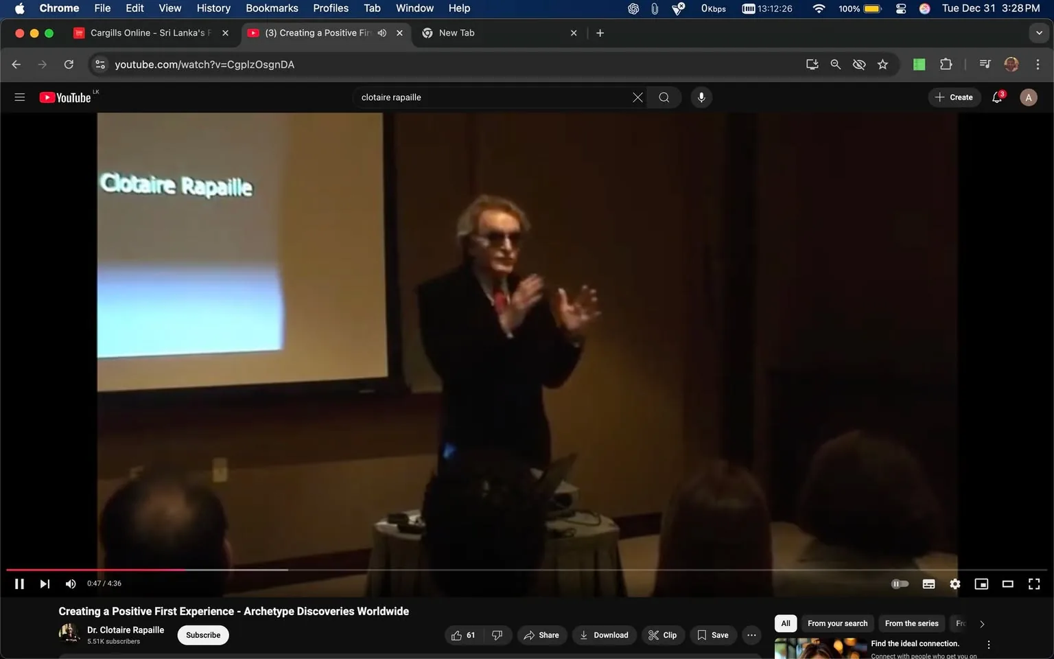 In the image, a speaker is presenting in front of an audience, gesturing with their hands as they speak. The individual is wearing sunglasses and a black suit, with a red tie, suggesting a formal presentation setting. Behind them, a screen displays the name "Clotaire Rapaille," indicating the speaker's identity. The audience appears to be seated, with some heads visible in the foreground, suggesting a small group attending the presentation. The environment looks like a conference room, with subdued lighting and a table in front of the speaker, possibly holding presentation materials. The overall atmosphere is professional and focused on the speaker's discourse.