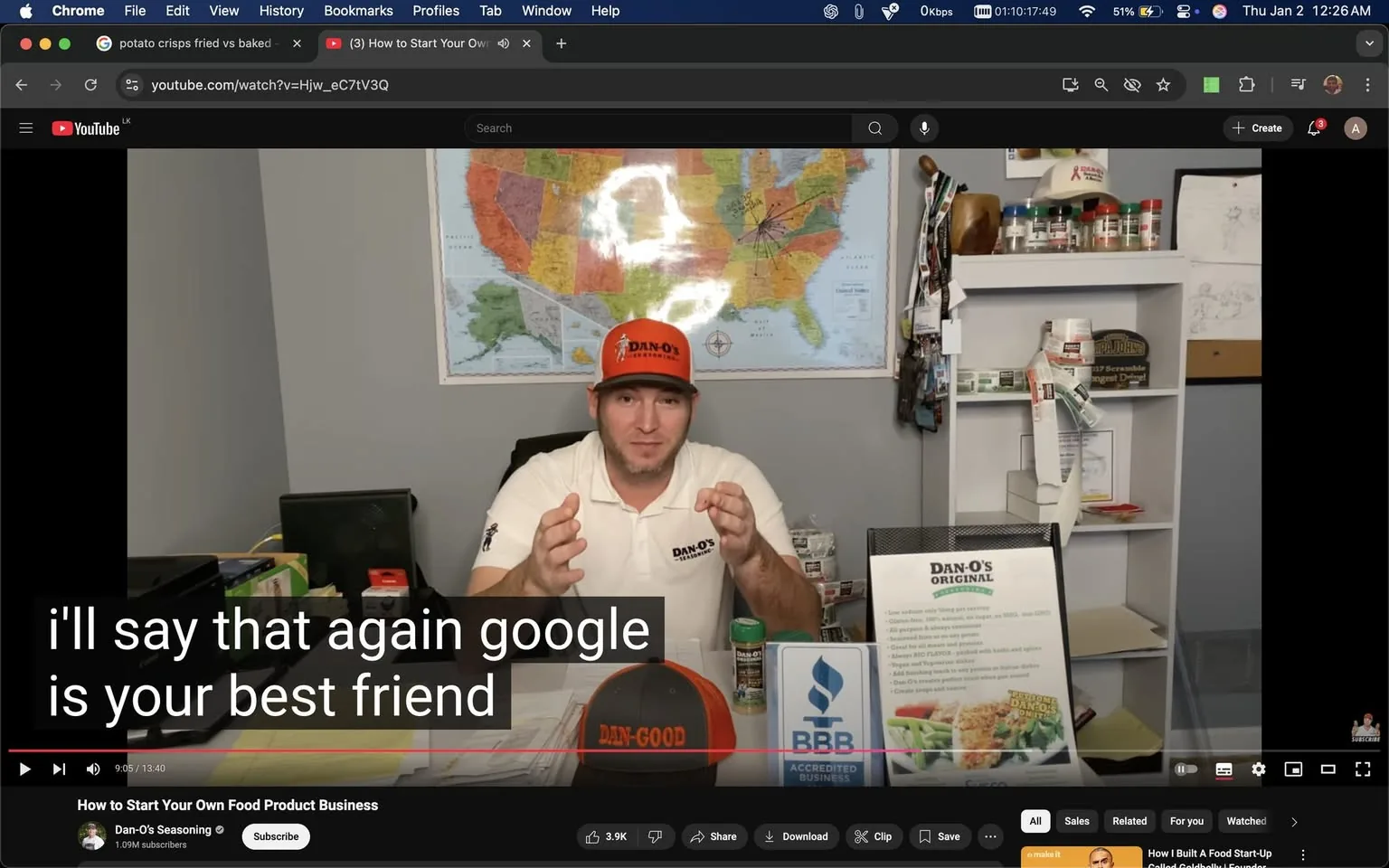 In the video, an unidentified adult male is seated at a desk, engaging with the audience about starting a food product business. He is wearing a white shirt with the "Dan-O's" logo and a matching orange cap that also features the brand name. The background includes a large map of the United States, and various items are visible on the desk, including a canister of seasoning, a brochure for Dan-O's, and a sign indicating BBB accreditation. The individual gestures with his hands while speaking, emphasizing his points. The setting appears to be an indoor office space, and the overall atmosphere is casual and informative.