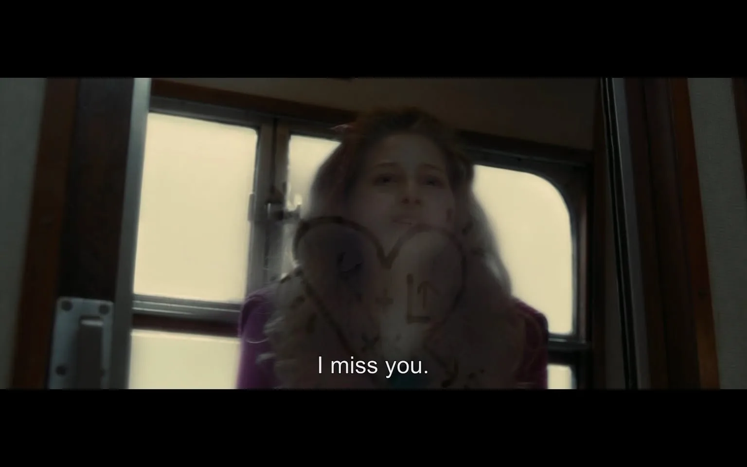 In the image, a young woman with long, wavy hair appears to be looking through a window, her expression conveying a sense of longing or sadness. She is dressed in a purple jacket, and the background suggests an indoor setting, possibly a train or bus, with foggy glass obscuring the view outside. On the glass, there is a heart shape drawn along with the text "I miss you," indicating a personal message. The overall mood is emotional, reflecting themes of separation or nostalgia.