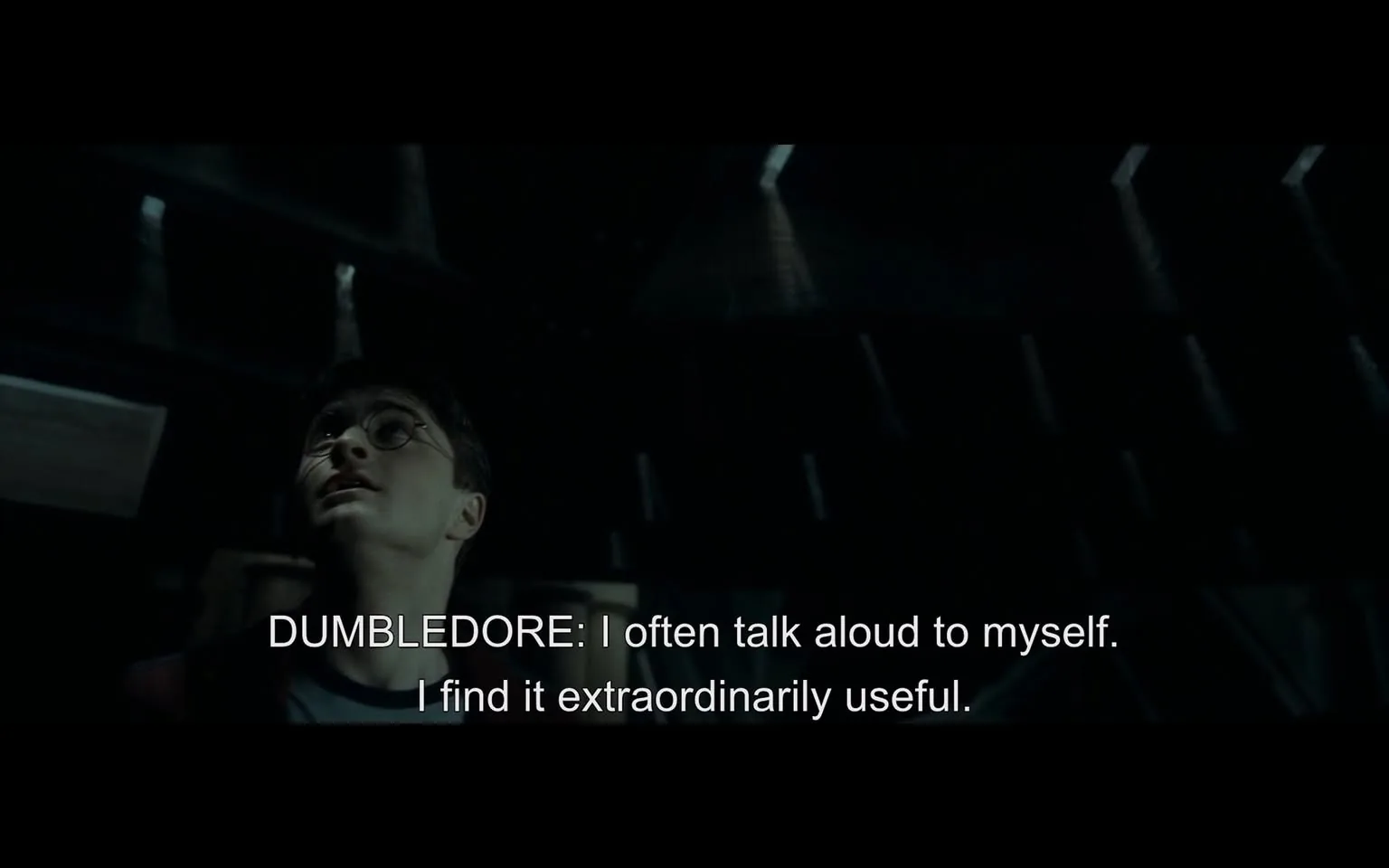 In a dimly lit setting, a character is looking upwards, appearing contemplative or engaged in thought. The character has short dark hair and wears round glasses, suggesting a youthful appearance. The background is shadowy, with faint light sources creating a dramatic atmosphere. The visible text indicates a quote from Dumbledore, stating, "I often talk aloud to myself. I find it extraordinarily useful." This line adds a layer of introspection to the scene. The overall mood is serious, reflecting a moment of reflection or wisdom.