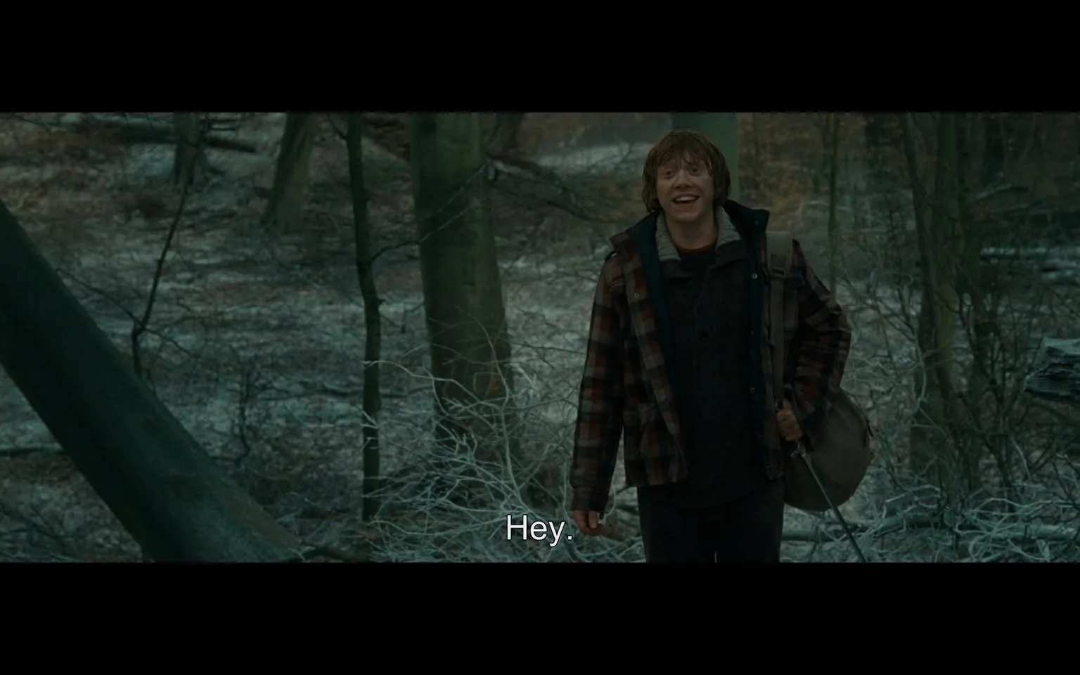 In the image, a young man stands in a forested area, smiling and greeting someone with a casual "Hey." He is wearing a plaid jacket over a dark sweater, and he carries a bag slung over his shoulder. The background features a mix of bare trees and underbrush, suggesting a cool or possibly early spring setting. The ground appears to be covered with a light layer of frost or snow, adding to the chilly atmosphere. The overall mood seems friendly and approachable, with the young man displaying an open expression.