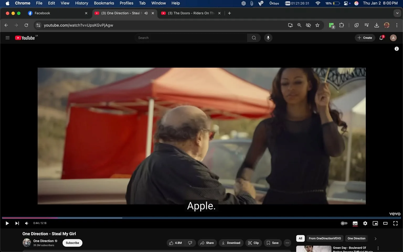 In the scene, an unidentified adult male is seated, appearing to engage with an unidentified adult female who stands beside him. The female is dressed in a black outfit and holds a decorative umbrella, while the male wears a black jacket and glasses. The background features a red tent, suggesting an outdoor setting, possibly a market or festival. The text "Apple" is visible, likely indicating a reference to the brand. The overall atmosphere seems casual, with a focus on the interaction between the two individuals.