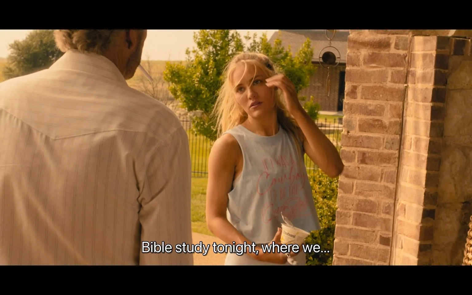 In the scene, an unidentified adult female is speaking to another individual, whose back is turned to the camera. The woman has long, blonde hair and is wearing a light blue sleeveless top with the text "If I was a cowboy, I'd be the..." partially visible. She appears to be in a casual outdoor setting, possibly at a home, with a well-maintained lawn and trees in the background. The time of day seems to be during daylight. The woman is holding a cup of Chobani yogurt, indicating a relaxed atmosphere. The dialogue suggests they are discussing a Bible study event planned for that evening.