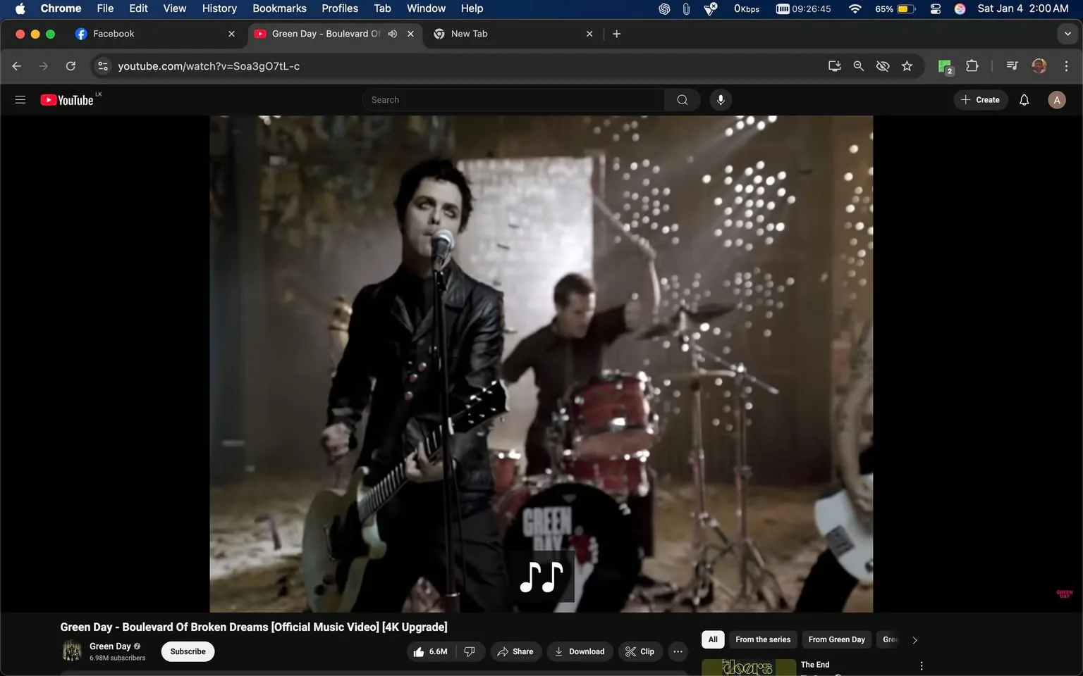 In the scene, a band is performing in a music video setting. The lead singer, dressed in a black leather jacket, is holding a guitar and singing into a microphone. Behind him, a drummer is playing a red drum set, contributing to the energetic atmosphere. The background features a textured wall with scattered lights creating a dynamic visual effect. The overall mood is intense and engaging, typical of a rock performance. The video is associated with the band Green Day, as indicated by the visible logo on the drum set and the title displayed on the screen. The setting appears to be indoors, likely in a studio designed for music videos.