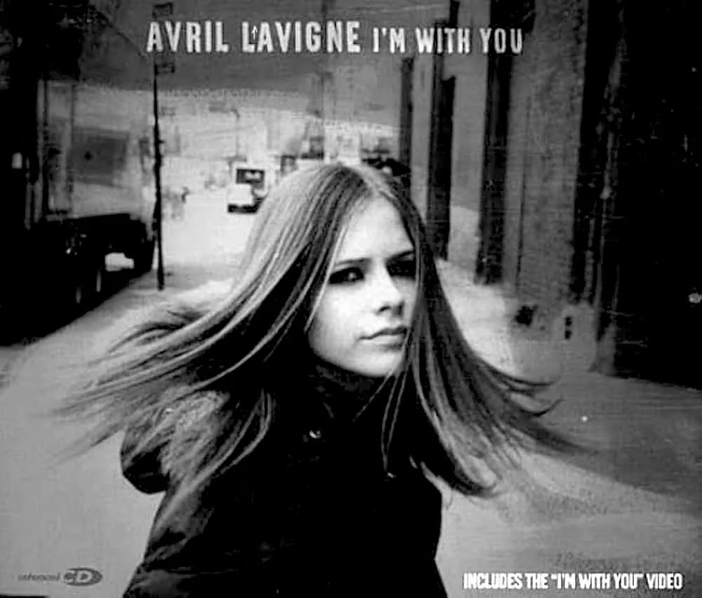 In the image, a young woman with long, flowing hair is captured in a black and white setting, giving it a nostalgic feel. She appears to be walking or turning, with her hair blowing slightly, suggesting movement. The background features urban elements, including buildings and street signs, which contribute to the overall atmosphere. Prominently displayed at the top is the text "AVRIL LAVIGNE I'M WITH YOU," indicating that this image is associated with a music title. The overall mood is reflective, possibly evoking themes of longing or searching. There are no visible brands or logos in the image.