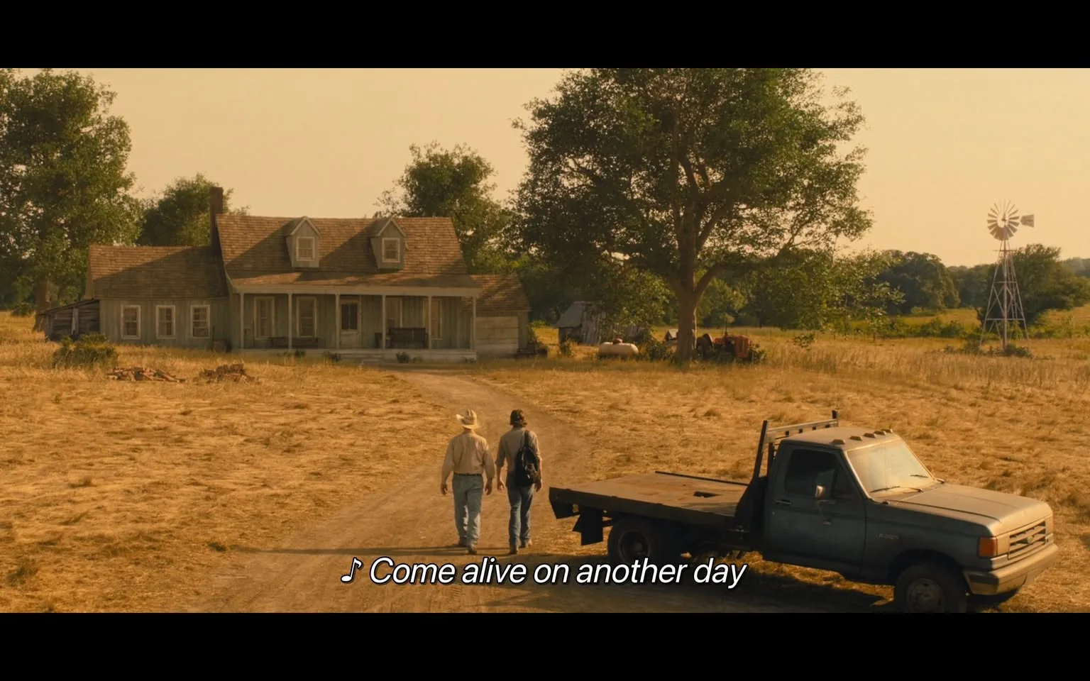 In a tranquil outdoor setting, two individuals are walking along a dirt path towards a large, two-story house with a porch. The house has a rustic appearance, featuring a gabled roof and multiple windows. Surrounding the house are tall trees and a field of dry grass, indicating a rural environment. A blue pickup truck is parked nearby, with a flatbed design, suggesting it may be used for work. In the background, a windmill can be seen, along with some farm equipment, enhancing the pastoral atmosphere. The sky appears warm and golden, hinting at either sunrise or sunset. The scene is accompanied by the lyrics "Come alive on another day," suggesting a hopeful or reflective mood.