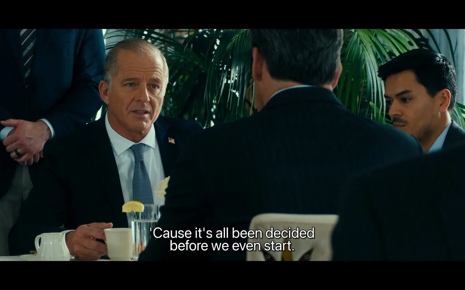 In a formal dining setting, an unidentified adult male in a suit is engaged in conversation with another man, whose back is visible. The first man has short, light-colored hair and is wearing a dark suit with a light blue tie, featuring a small flag pin on his lapel. He appears to be making a serious point, stating, "'Cause it's all been decided before we even start." The background includes lush green plants, suggesting an indoor environment, likely a restaurant or a business meeting space. Another unidentified adult male is partially visible, listening intently. The atmosphere seems tense, indicating a significant discussion taking place.