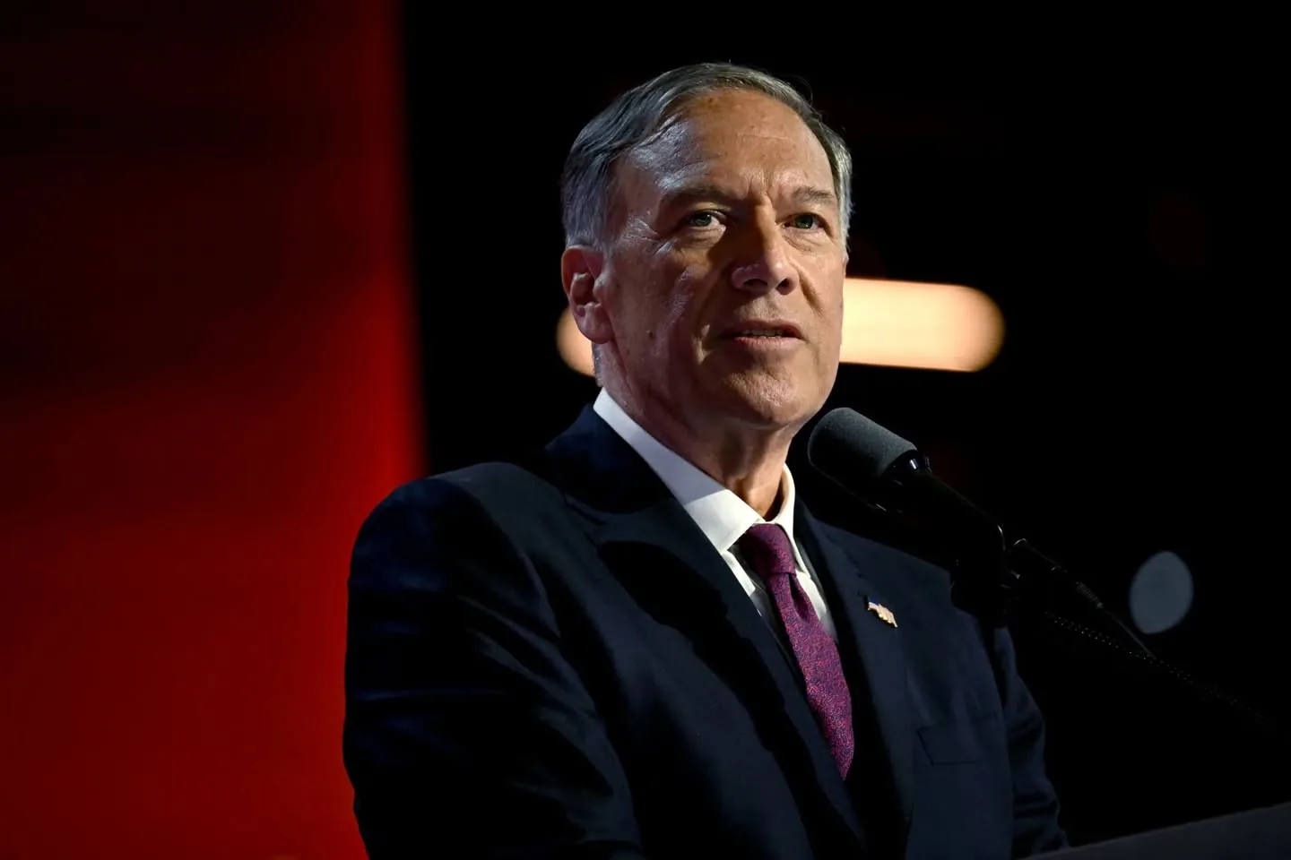 In the image, a man is speaking at a podium, appearing to address an audience. He is dressed in a dark suit with a white shirt and a patterned tie, which is a deep shade of purple. His hair is gray and neatly styled. The background features a gradient of red and dark tones, suggesting a formal or significant event. The man has a serious expression, indicating he is likely delivering an important message. A microphone is positioned in front of him, and there are soft lights illuminating the scene, enhancing the formal atmosphere. No visible brands or logos are present in the image.