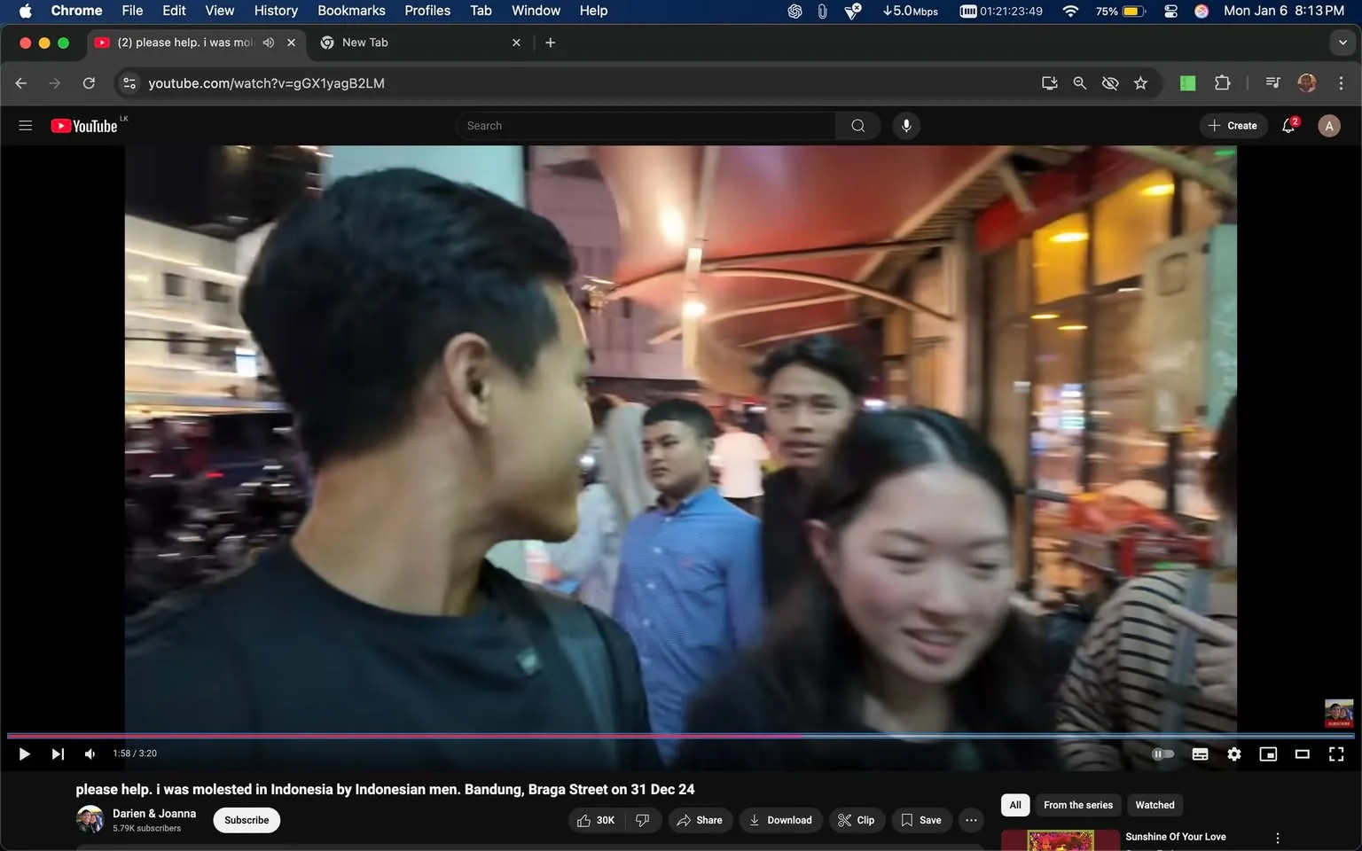 In a bustling street scene, a group of individuals is gathered, likely in an urban area during the evening. The main focus is on a young man with short black hair, wearing a black shirt, who is turned slightly to the left, engaging with a young woman beside him. She has long dark hair and is wearing a black top. In the background, several other individuals can be seen, including a man in a blue shirt and another person with a striped shirt. The setting appears lively, with illuminated storefronts and a canopy overhead, suggesting a vibrant atmosphere typical of a night market or street fair. The expressions of the people are varied, with some appearing engaged in conversation while others look on. No visible brands or logos are present in the image.