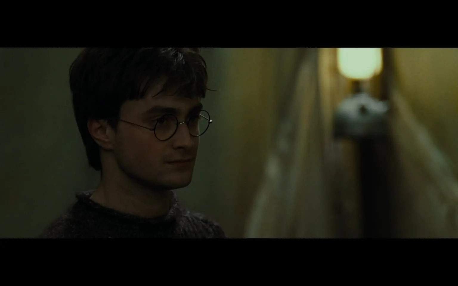 In a dimly lit corridor, a young man with short, dark hair and round glasses stands in profile, appearing contemplative or serious. He wears a dark, knitted sweater, and the background is blurred, emphasizing his presence. The walls of the corridor are a muted color, possibly greenish, and there is a soft light source visible in the background, creating a moody atmosphere. The overall setting suggests a moment of introspection or tension, typical of a narrative scene. No identifiable brands or logos are present in the image.