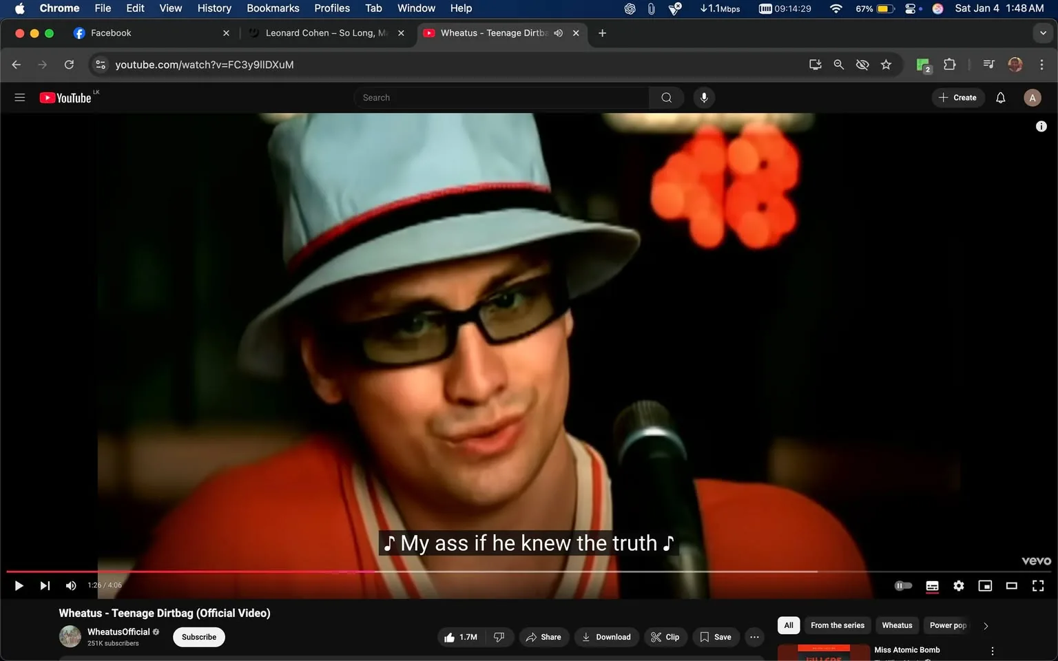 In the image, a person is performing in what appears to be a music video. They are wearing a light blue hat with a red band and dark sunglasses. Their expression is somewhat playful as they sing into a microphone. The background is dimly lit, with soft, out-of-focus lights creating a warm ambiance. The lyrics displayed on the screen read, "My ass if he knew the truth," suggesting a narrative element to the performance. The overall setting seems to be indoors, likely in a music venue or studio, contributing to the intimate atmosphere of the scene.