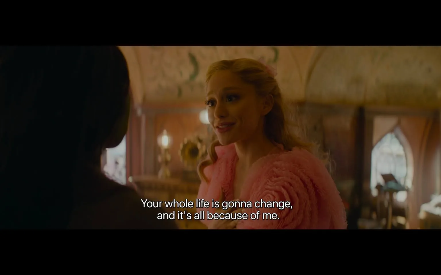 In the scene, a character with long, wavy hair styled in soft curls is wearing a fluffy pink garment, possibly a coat or shawl. She has a bright smile and is engaged in conversation, conveying a sense of excitement or confidence. The background features ornate decor, including intricate patterns on the walls and warm lighting from lamps, suggesting an elegant indoor setting. The character is speaking to another individual, whose face is partially visible, indicating a moment of interaction. The dialogue displayed on the screen reads, "Your whole life is gonna change, and it's all because of me," hinting at a significant plot development.