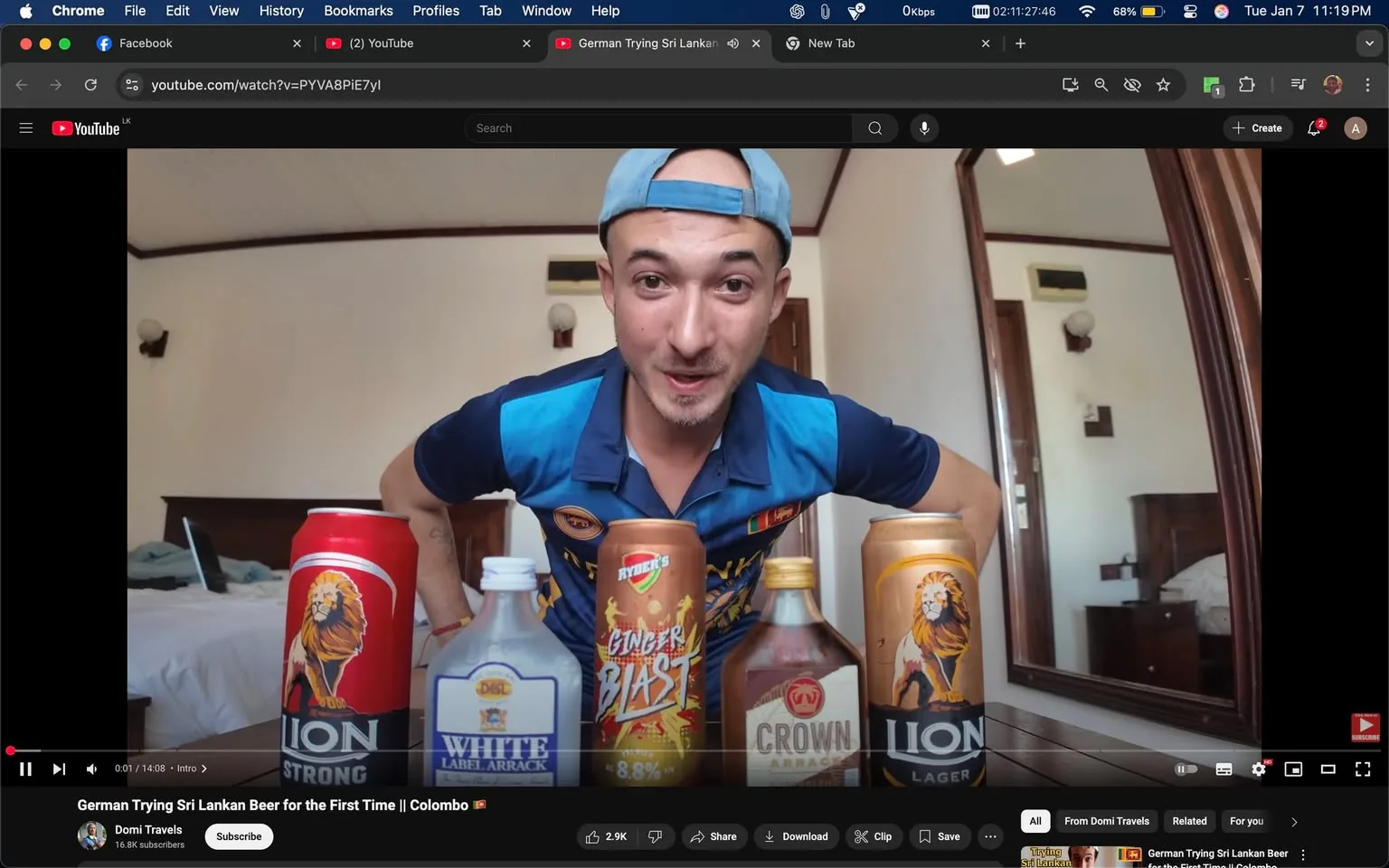 An unidentified adult male is positioned in front of a table displaying several alcoholic beverages, including cans and bottles. The drinks include "Lion Strong," "White Label Arrack," "Crown Arrack," and "Ryder's Ginger Blast." The individual is wearing a blue polo shirt with a logo and a light blue cap, leaning forward with a playful expression. The setting appears to be indoors, likely in a hotel room, with a mirror reflecting part of the room in the background. The atmosphere suggests a casual or entertaining context, possibly related to tasting or reviewing the drinks.
