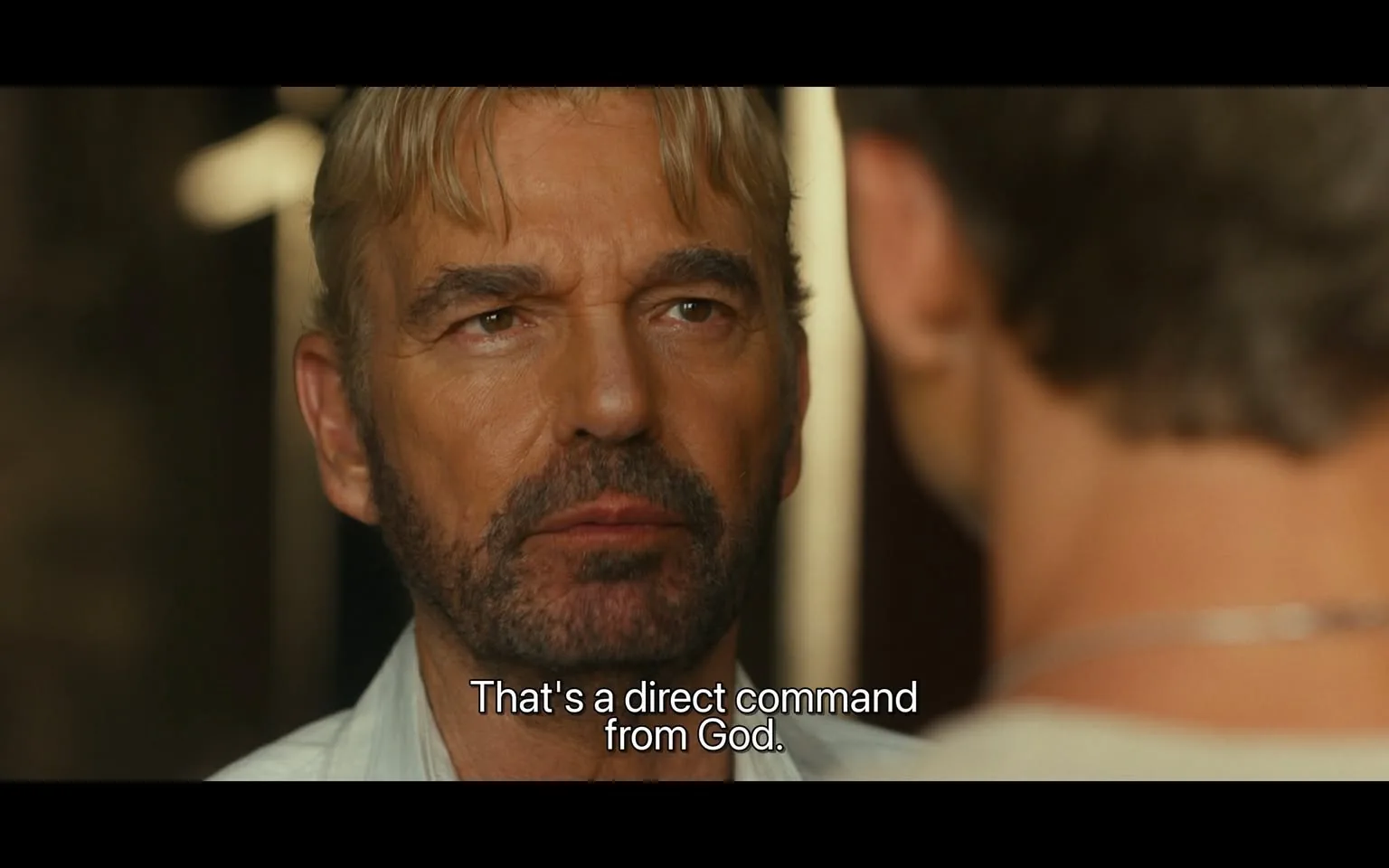 In the image, an unidentified adult male with light blonde hair and a beard is engaged in a serious conversation. His expression is intense, and he appears to be making a significant statement, as indicated by the text overlay that reads, "That's a direct command from God." The background suggests an indoor setting, with soft lighting that creates a dramatic atmosphere. The other individual in the frame is partially visible, but their features are not clear. The focus is on the man speaking, who is wearing a light-colored shirt, and his demeanor conveys a sense of authority or urgency.