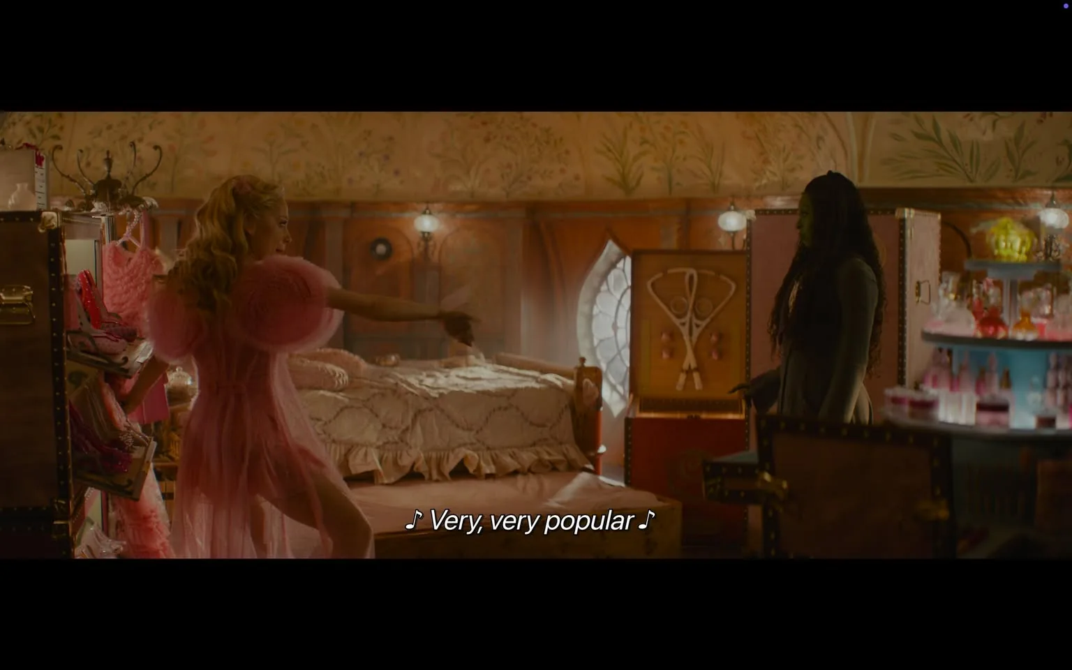 In a vibrant, whimsical room, two characters are engaged in a lively interaction. One character, dressed in a fluffy pink outfit, is striking a pose and appears to be singing or performing, with the lyrics "Very, very popular" displayed on the screen. The other character, dressed in darker clothing, stands nearby, seemingly observing the performance. The room is decorated with floral wallpaper and features a bed with a quilted cover, a wooden chest with decorative elements, and shelves filled with colorful bottles, suggesting a playful and imaginative atmosphere. The lighting is warm, enhancing the cozy feel of the space.