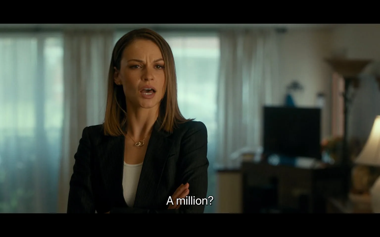 In the scene, an unidentified adult woman stands with her arms crossed, displaying a look of surprise or disbelief as she speaks the words, "A million?" Her expression suggests she is engaged in a serious conversation. She is dressed in a dark blazer over a white top, and her hair is styled in loose waves. The background features a well-lit indoor setting with sheer curtains, a dark piece of furniture, and a television, indicating a casual yet professional environment. The lighting is soft, suggesting it could be daytime.