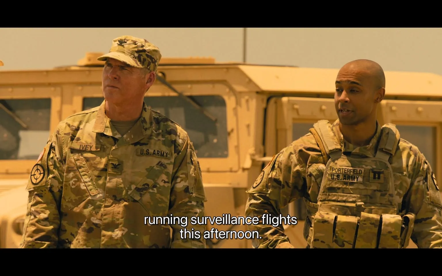 In the scene, two soldiers are present, both wearing U.S. Army camouflage uniforms. The soldier on the left has the last name "Ivey" visible on his uniform, while the soldier on the right has "Porterfield" displayed. They appear to be engaged in a conversation about running surveillance flights later in the day. The background features military vehicles, specifically a Humvee, indicating an outdoor setting likely in a desert or arid environment. The atmosphere seems serious, reflecting their military context. The soldiers' expressions are focused, suggesting they are discussing important operational details.