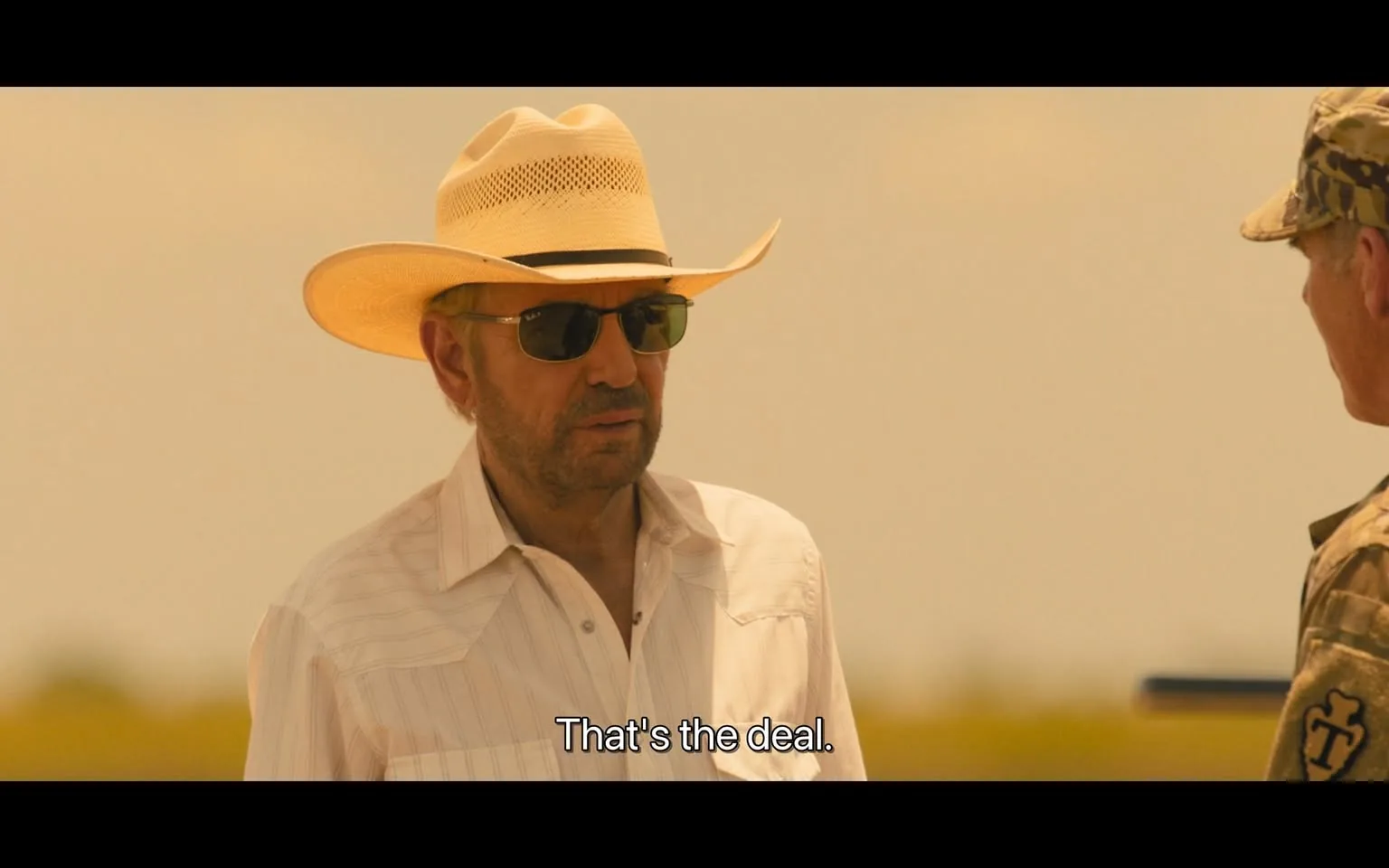 In the scene, an unidentified adult man is wearing a white, short-sleeved shirt and a wide-brimmed straw hat, complemented by dark sunglasses. He appears to be engaged in a serious conversation, stating, "That's the deal." The background is hazy, suggesting an outdoor setting, possibly under bright sunlight. Another individual, dressed in military attire, stands nearby, listening intently. The overall atmosphere conveys a tense negotiation or agreement. There are no visible weapons or brands in the image.