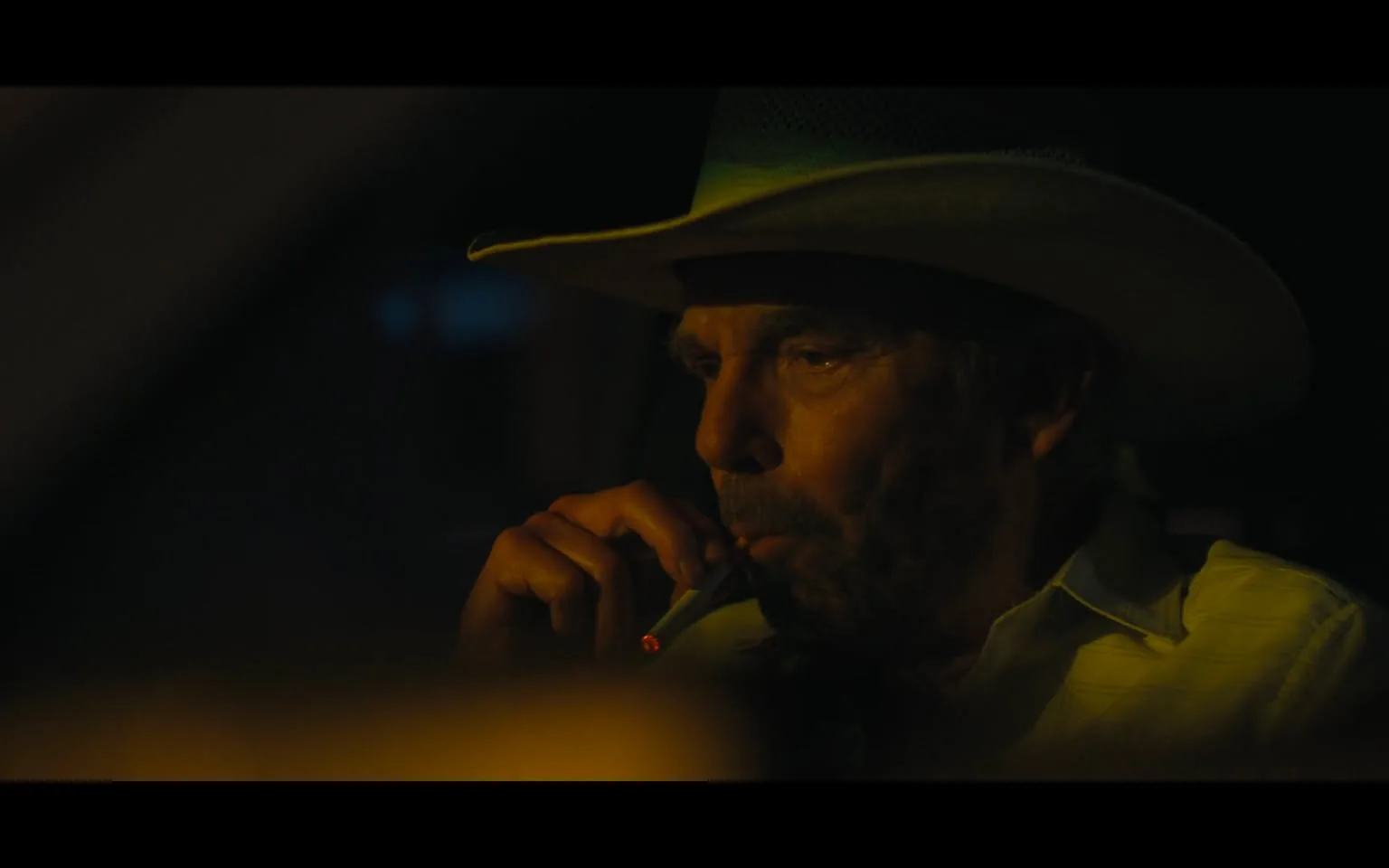 An adult male is depicted in a dimly lit setting, possibly inside a vehicle, as he smokes a cigarette. He wears a wide-brimmed cowboy hat and a light-colored shirt, which has a subtle pattern. His expression appears serious and contemplative, with visible signs of age, such as wrinkles and a rugged demeanor. The background is dark, with hints of blue light suggesting nighttime. There are no visible brands or logos in the image, and no elements indicating violence or sexual content. The overall atmosphere conveys a sense of tension or introspection.