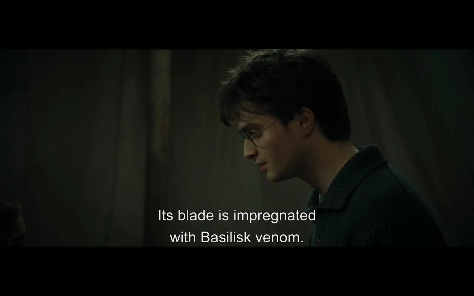 In the scene, a young man with glasses is engaged in a serious conversation, his expression focused and contemplative. He has tousled dark hair and is wearing a dark sweater with a ribbed collar. The background is dimly lit, suggesting an indoor setting that feels tense or dramatic. The dialogue displayed on the screen mentions a blade that is "impregnated with Basilisk venom," indicating the weapon's dangerous nature. The atmosphere is charged with a sense of urgency or importance, likely related to a larger narrative involving magic or fantasy elements.