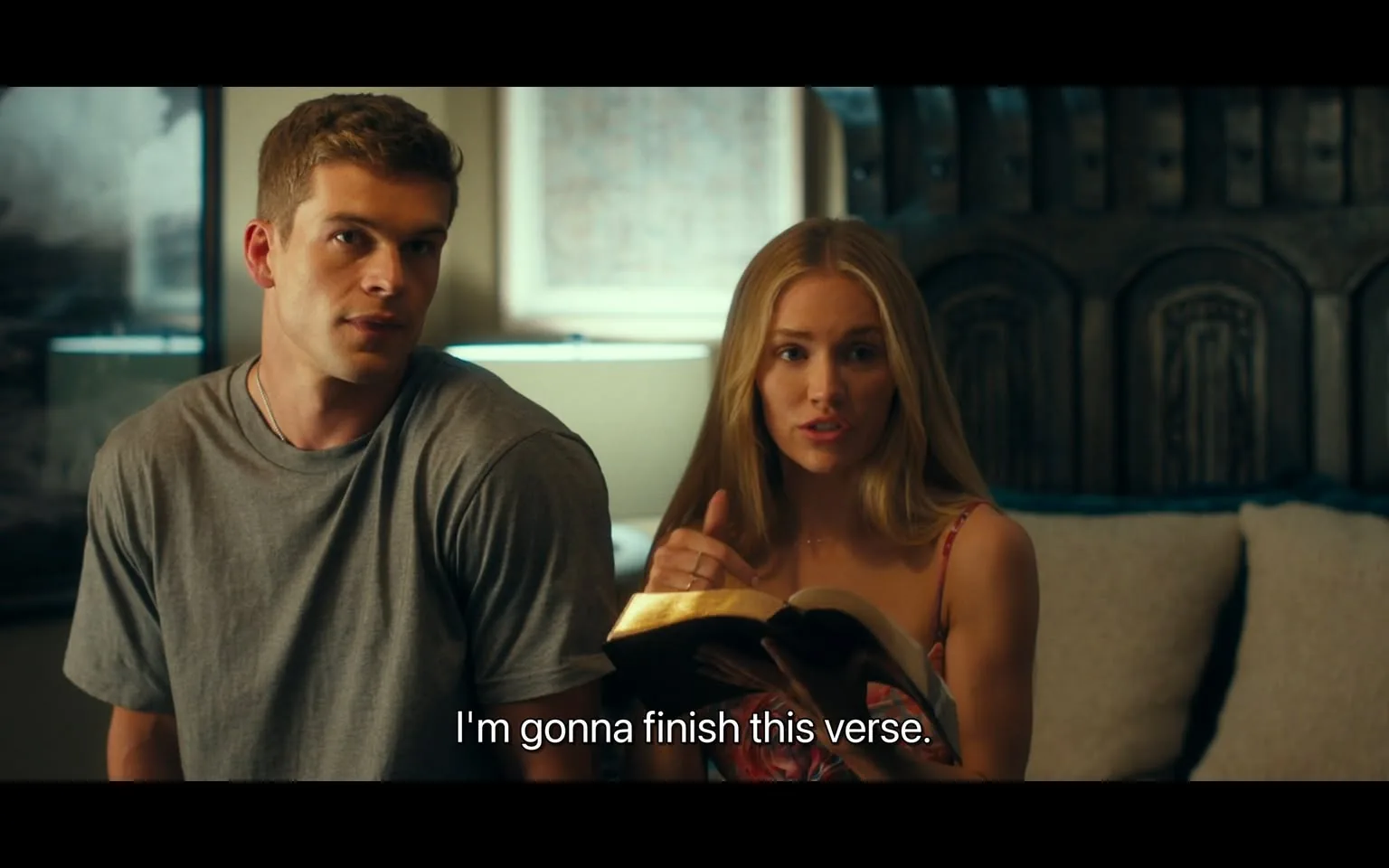In the scene, two individuals are seated indoors, engaged in a conversation. One person, a young adult male, is wearing a gray t-shirt and has a neutral expression as he listens. The other person, a young adult female, is holding an open book, likely a Bible, and appears animated as she speaks, stating, "I'm gonna finish this verse." She has long, straight blonde hair and is dressed in a colorful, patterned outfit. The background features a softly lit room with a couch and decorative elements, suggesting a casual and intimate setting. The lighting is warm, indicating it could be evening.