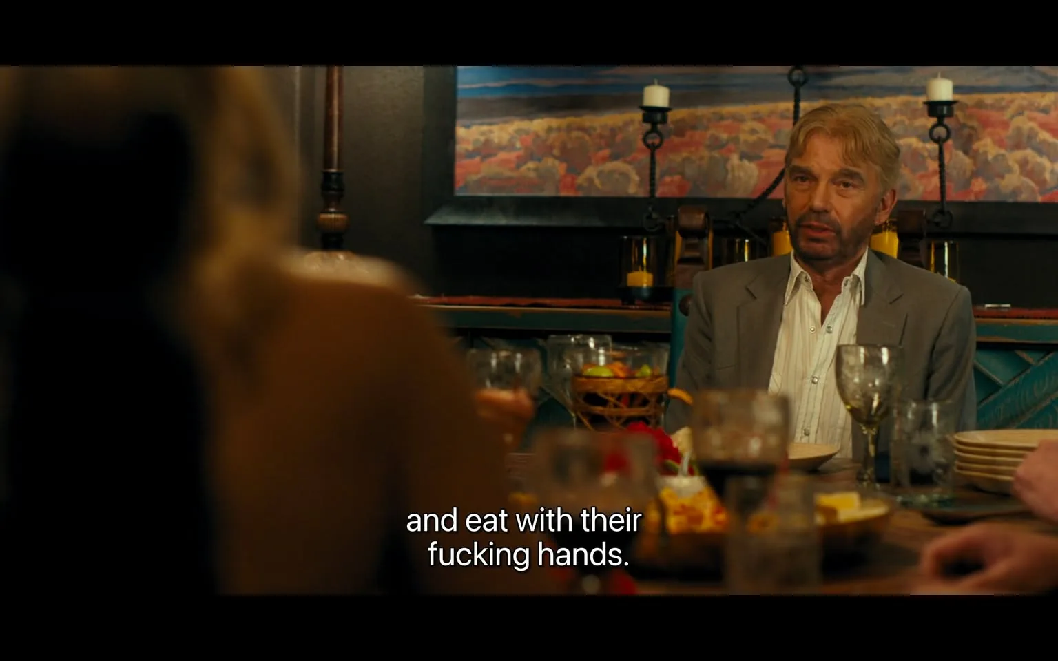 In the scene, an unidentified adult male is seated at a dining table, dressed in a light gray suit with a white shirt. He appears to be engaged in a heated discussion, expressing frustration about people eating with their hands. The table is set with various dishes, including what looks like a colorful assortment of food, and several glasses filled with drinks. The setting is indoors, with a warm ambiance created by candlelight and a painting of a landscape in the background. The atmosphere suggests a social gathering, but the tone of the conversation is intense, highlighted by the use of strong language. Other individuals are present, but their faces are not clearly visible due to the angle of the shot.