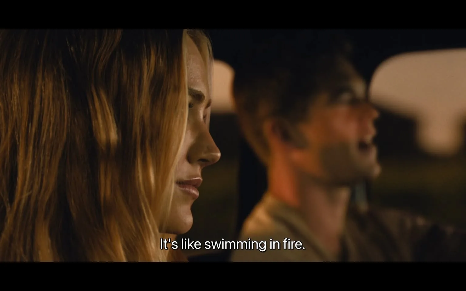 In the image, two individuals are seated inside a car, with the focus primarily on a young woman with long, wavy hair. She appears to be listening intently, her expression thoughtful. The young man next to her is engaged in conversation, speaking animatedly. The lighting suggests it is either dusk or dawn, creating a warm ambiance inside the vehicle. The dialogue displayed on the screen reads, "It's like swimming in fire," indicating an emotional or intense topic of discussion. The background is blurred, emphasizing the characters and their interaction. No brands or logos are visible in the image.