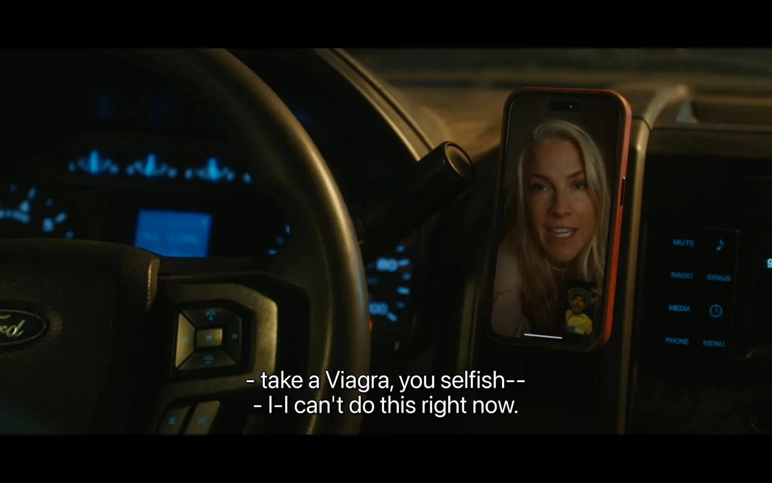 In the scene, an unidentified adult is seen speaking on a phone, which is mounted on the dashboard of a vehicle, specifically a Ford. The individual on the phone has long, light-colored hair and appears to be expressing frustration or urgency, as indicated by their facial expression and tone. The dialogue includes phrases like "take a Viagra, you selfish" and "I can't do this right now," suggesting a personal and possibly heated conversation. The vehicle's dashboard displays blue-lit indicators, and the phone screen shows a small image of another person, possibly a child or a character, indicating a casual or familial context. The setting appears to be nighttime, given the lighting and the vehicle's interior ambiance.