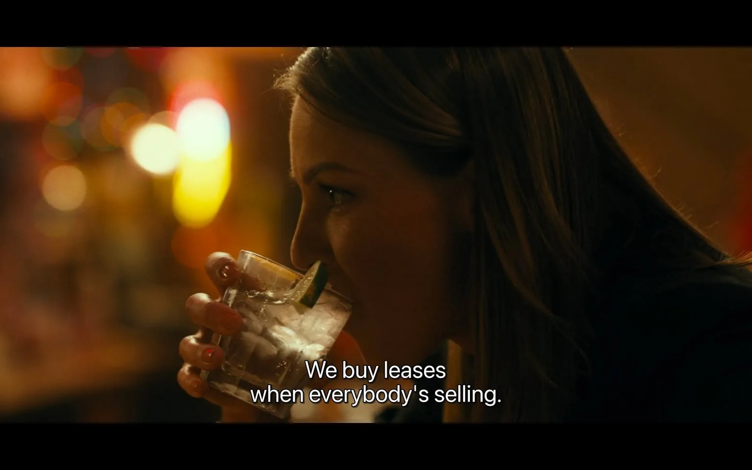 In the image, an unidentified adult is seen holding a glass with ice and a slice of lime, suggesting they are enjoying a drink in a social setting, likely a bar or restaurant. The background is softly illuminated with colorful bokeh lights, creating a warm and inviting atmosphere. The person appears to be engaged in conversation, as indicated by the text overlay that reads, "We buy leases when everybody's selling." Their expression is focused, hinting at a serious discussion. The overall ambiance suggests a casual yet intimate gathering.