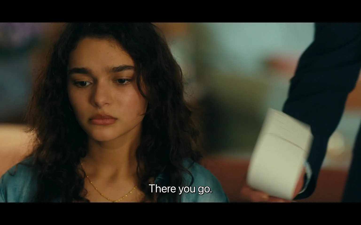 In the scene, a young woman with curly hair and a neutral expression appears to be in a moment of contemplation or concern. She is wearing a light blue garment and has a delicate necklace. The background suggests an indoor setting, possibly a casual or intimate environment, with soft lighting. A hand, presumably belonging to another person, is reaching out with a small white object, possibly a piece of paper or a note, while the dialogue reads, "There you go." The overall mood seems to convey a sense of seriousness or emotional weight.