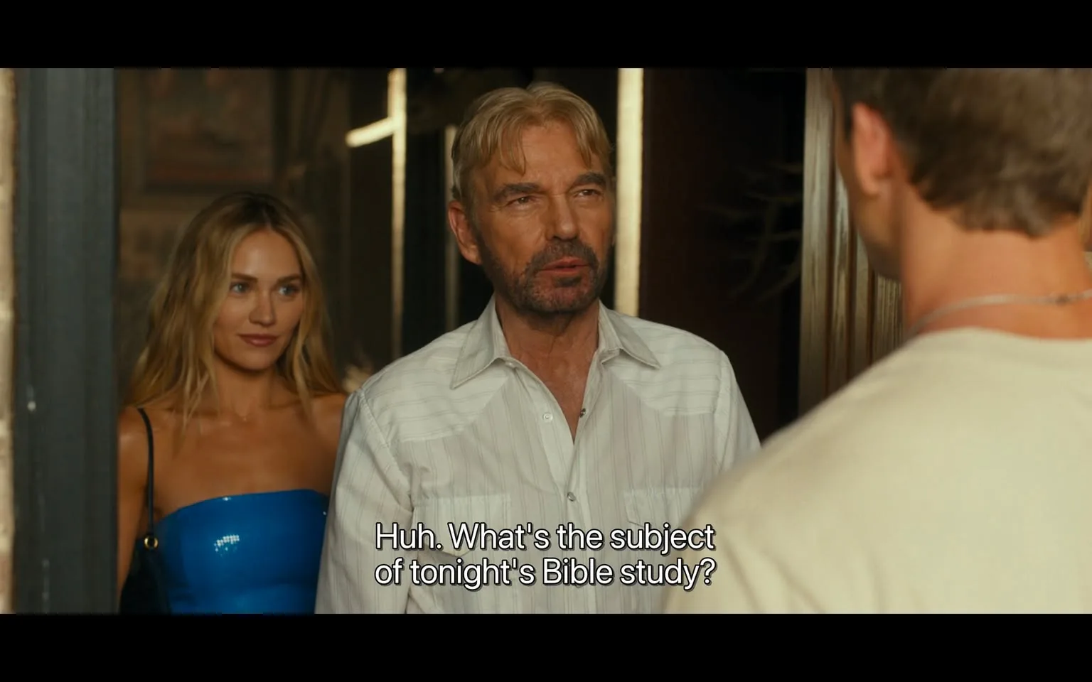 In the scene, two adults are engaged in a conversation at a doorway. One adult, a man with light-colored hair and a beard, is wearing a white button-up shirt with vertical stripes. He appears to be in his late 40s to early 60s. The other adult, a woman with long, wavy hair, is dressed in a form-fitting blue dress and stands slightly behind him, observing the interaction. The man is asking a question about a Bible study, suggesting a casual or humorous tone. The setting appears to be indoors, with wooden elements visible in the background, indicating a relaxed atmosphere. The lighting is warm, suggesting it could be evening.