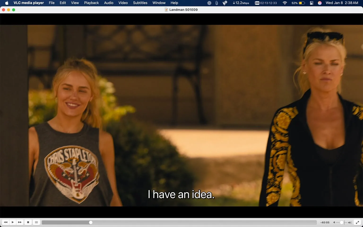 In the scene, two individuals are present outdoors, with a warm, sunny atmosphere suggesting daytime. The person on the left, wearing a sleeveless gray shirt with a graphic featuring a tiger and the name "Chris Stapleton," appears to be smiling and is engaged in conversation. The other individual, on the right, is dressed in a black jacket adorned with yellow floral patterns and has a serious expression. The background features a blurred view of greenery and a structure, indicating a residential or casual setting. The subtitle reads, "I have an idea," suggesting a moment of inspiration or planning.