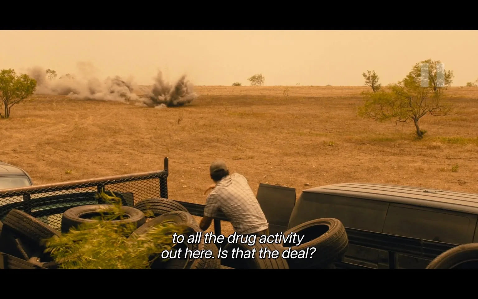 In the scene, an unidentified adult is positioned near a vehicle, looking towards a distant explosion that has just occurred in a dry, open landscape. The ground is barren, with sparse vegetation and a few trees scattered in the background. The adult is wearing a light-colored, checkered shirt and appears to be in a state of alertness or concern, suggesting a tense situation. The explosion creates a cloud of dust and debris, indicating possible violence or conflict related to drug activity, as referenced in the dialogue. The setting is outdoors, likely during the day, given the visibility of the explosion and the clear sky. The presence of tires piled near the vehicle adds to the rugged, makeshift environment.