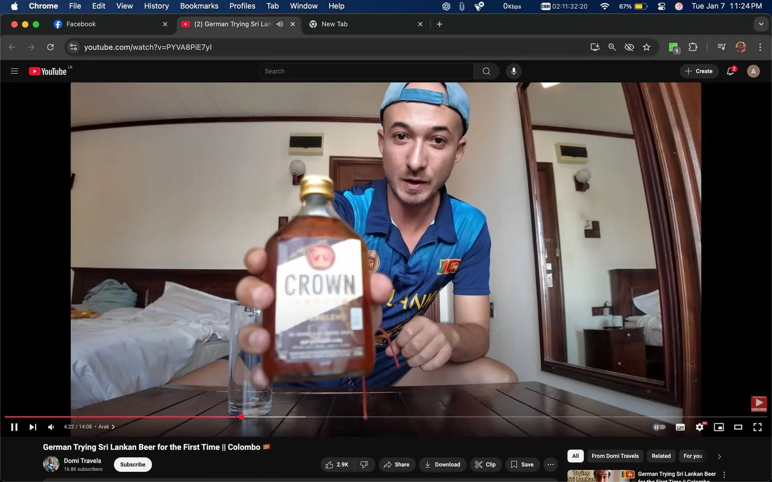 In the image, an unidentified adult male is seated at a wooden table in a hotel room, holding a bottle labeled "CROWN." He is wearing a blue sports shirt with a Sri Lankan flag emblem and a light blue cap. The background reveals a bed with rumpled sheets and a wooden nightstand. A glass is placed on the table next to him. The setting appears casual, likely during the daytime, as indicated by the natural light coming through the room. The focus is on the bottle, suggesting a theme related to trying or tasting the beverage.