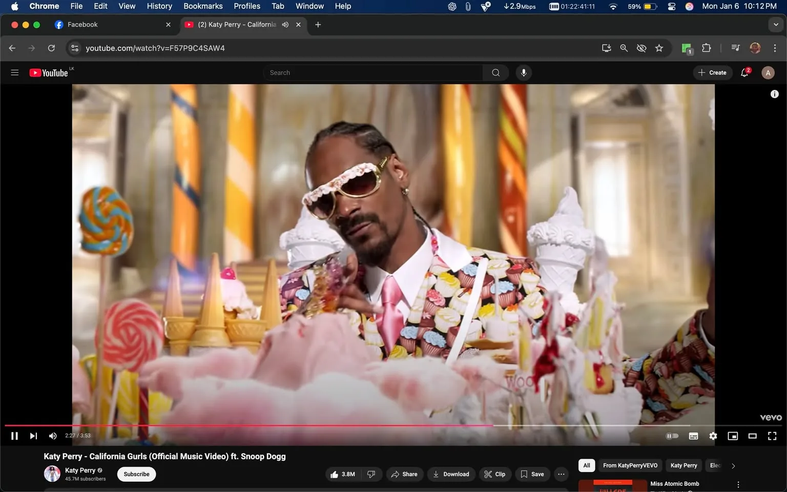 In the image, a man is seated at a lavish table adorned with various sweets, including ice cream cones, cotton candy, and large lollipops. He wears a vibrant suit decorated with cupcake patterns and oversized sunglasses. The setting appears to be whimsical, resembling a candy-themed environment, with bright colors and ornate decorations in the background. The man has a relaxed expression, seemingly enjoying the playful atmosphere. The scene is lively and colorful, emphasizing a fun, indulgent theme.