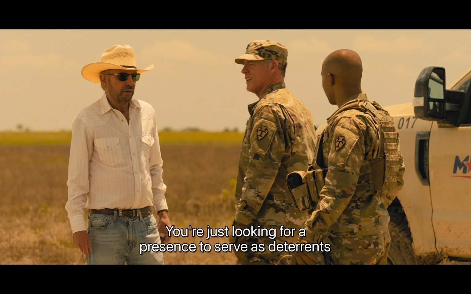In the scene, a civilian man wearing a white button-up shirt and a wide-brimmed straw hat stands in a field, engaged in conversation with two military personnel. The civilian appears to be in his late 50s to early 60s, with sunglasses and a serious expression. The military personnel are dressed in camouflage uniforms, one of whom is smiling, suggesting a friendly exchange. The background features a vast, open field under a clear sky, indicating a rural setting. The text overlay reads, "You're just looking for a presence to serve as deterrents," hinting at a discussion about security or military presence. No weapons or explicit content are visible in the image.