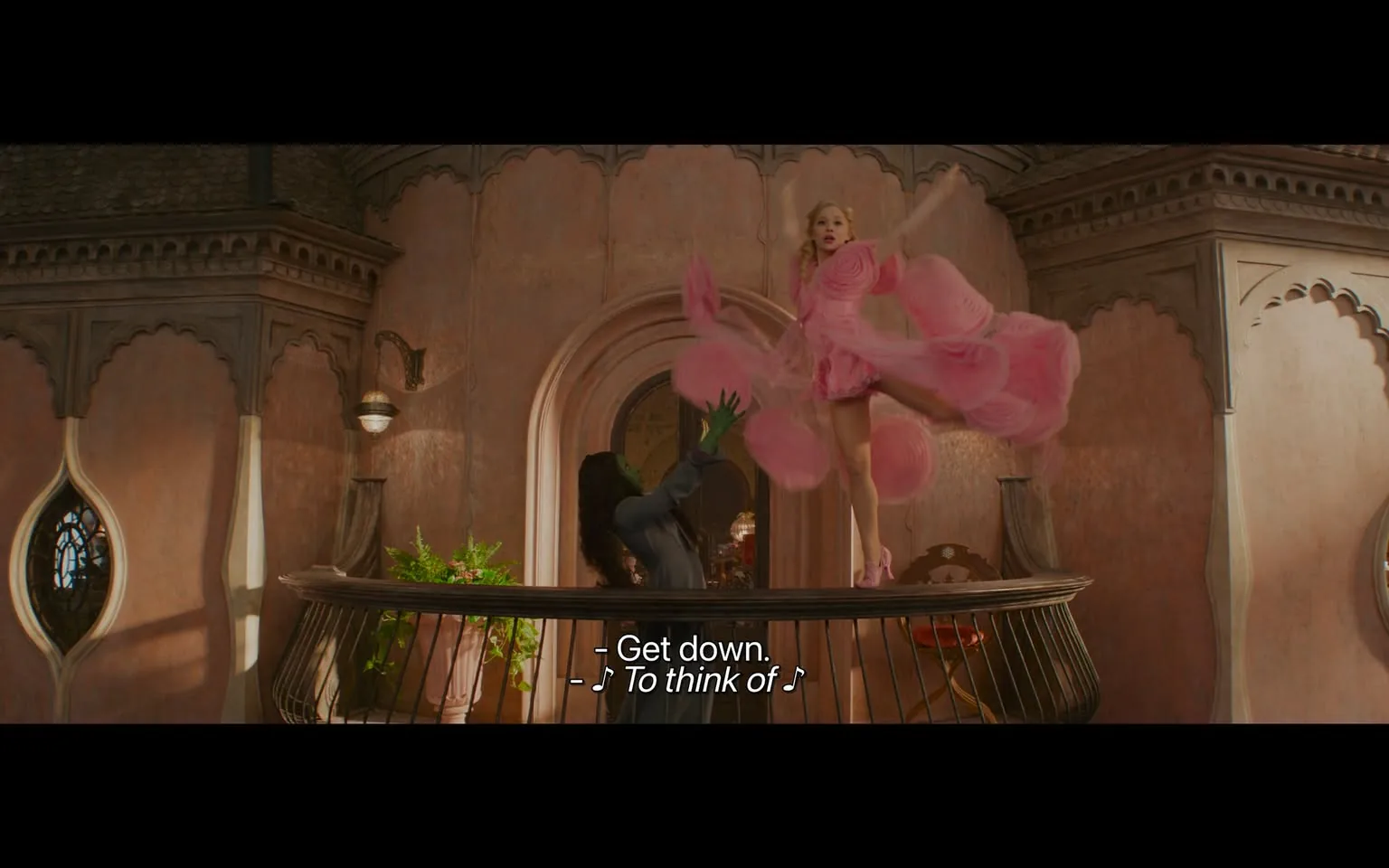 In a vibrant indoor setting, a character dressed in a voluminous pink dress is striking a dynamic pose, seemingly leaping or dancing above a balcony. The dress features large, circular embellishments that add to its dramatic flair. Below, another character, dressed in dark clothing with green skin, appears to be reaching up towards the pink-dressed figure, possibly in a gesture of surprise or urgency. The background features ornate architectural details with arched windows and a warm, pinkish wall, suggesting a whimsical or fantastical environment. The scene is lively, with the characters engaged in a moment that conveys energy and emotion, accompanied by the text "Get down" and musical notes, indicating a performance or musical context.