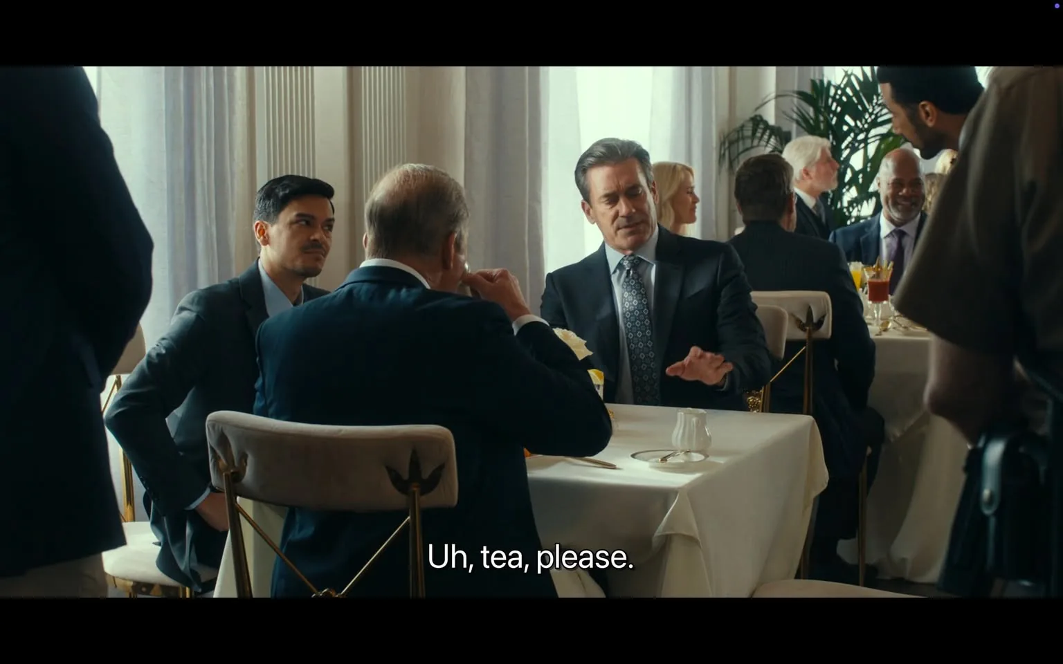 In a well-lit dining area, a group of individuals is seated at a table, engaged in conversation. The main focus is on a man in a suit, gesturing with his hands as he speaks. He has short, styled hair and appears to be in his 40s. To his left, another man, younger with dark hair, listens attentively. A third man, older and balding, is seated across from them, holding a drink and looking thoughtful. The table is elegantly set with white tablecloths and a small vase with a flower. In the background, other diners can be seen, and there are large windows allowing natural light to fill the space. The atmosphere seems formal and social, likely during a business or networking event. The dialogue includes a request for tea, indicating a casual yet professional interaction.