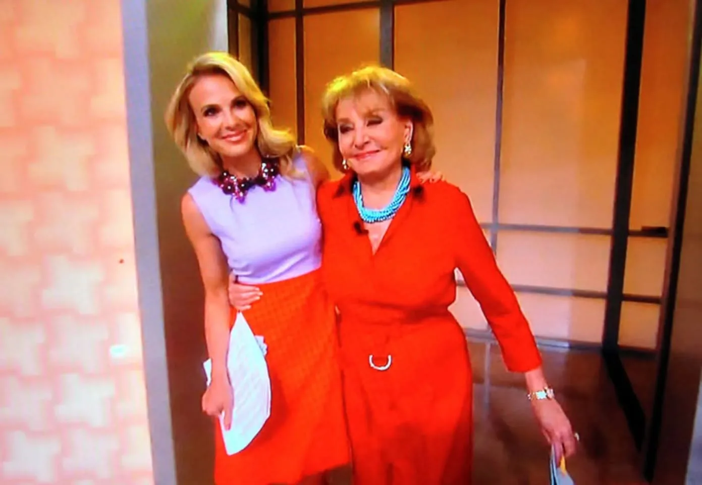 In the image, two adults are standing closely together, both smiling warmly. One is wearing a light purple top with a colorful necklace and an orange skirt, while the other is dressed in a bright red outfit with a belt and turquoise jewelry. They appear to be in a cheerful mood, possibly celebrating or concluding an event. The background features a softly lit indoor setting with a neutral-colored wall and a simple structure, suggesting a studio or stage environment. Both individuals are holding papers, indicating they may have just finished a presentation or segment.