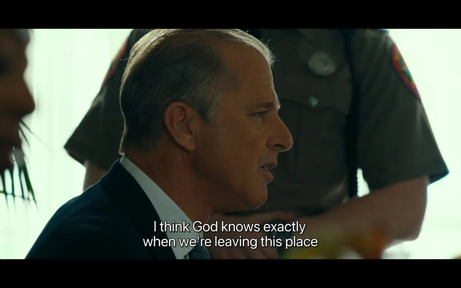 In the scene, an unidentified adult male is engaged in a serious conversation, expressing a belief that "God knows exactly when we're leaving this place." His expression appears thoughtful and somewhat tense, indicating the gravity of the discussion. He is dressed in a formal suit, with neatly combed hair. In the background, a law enforcement officer is partially visible, suggesting a formal or official setting. The environment appears to be indoors, with natural light filtering through a window, creating a somewhat bright atmosphere. The presence of a police officer hints at a serious context, possibly involving legal or law enforcement matters.