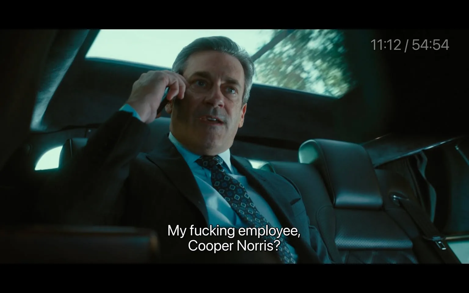 In the scene, a man in a suit is seated in a vehicle, appearing to be in a conversation on his phone. He has short, neatly styled hair and is wearing a tie with a geometric pattern. The interior of the car is dark, with leather seats visible. The man looks frustrated as he speaks, saying, "My fucking employee, Cooper Norris?" The background shows a blurred view of trees, indicating that the vehicle is in motion. The overall atmosphere suggests a tense moment in a professional context. There are no visible brands or logos in the image.