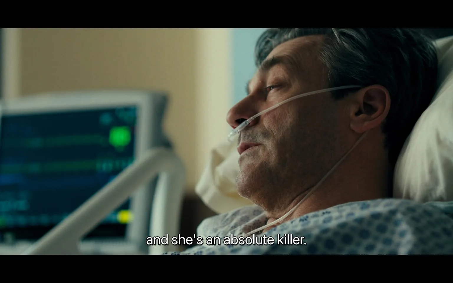 In a hospital room, an unidentified adult male lies in a hospital bed, appearing to be in a serious condition. He has a nasal cannula for oxygen support and is dressed in a hospital gown with a blue and white pattern. The background features a medical monitor displaying green and yellow lines, likely indicating vital signs. The man speaks with a serious expression, mentioning someone as "an absolute killer," suggesting a tense or dramatic conversation. The setting is well-lit, typical of a hospital environment, and conveys a sense of urgency or concern.