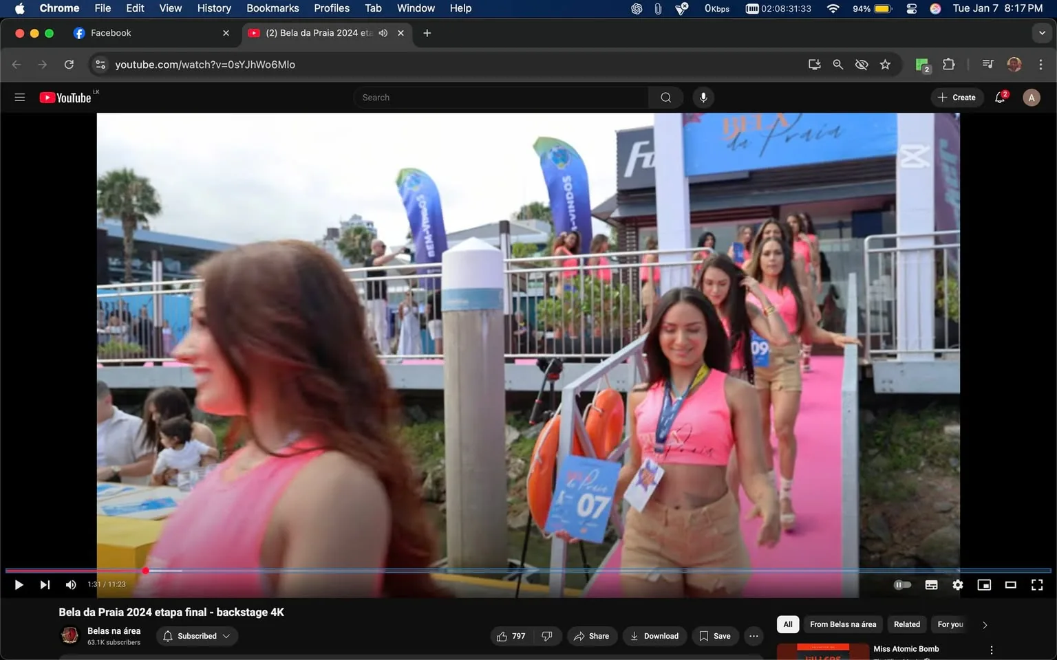 In a vibrant outdoor setting, several participants are seen walking along a pink runway, likely as part of a beauty pageant event titled "Bela da Praia 2024." The women are dressed in matching pink tops and shorts, showcasing a coordinated look. Each participant carries a sign with a number, indicating their order or identification for the event. The background features a modern structure with a sign displaying the event name, along with colorful flags fluttering in the breeze. The atmosphere appears lively, with onlookers present, suggesting a festive occasion. The scene captures a moment of anticipation as the participants prepare to showcase themselves.