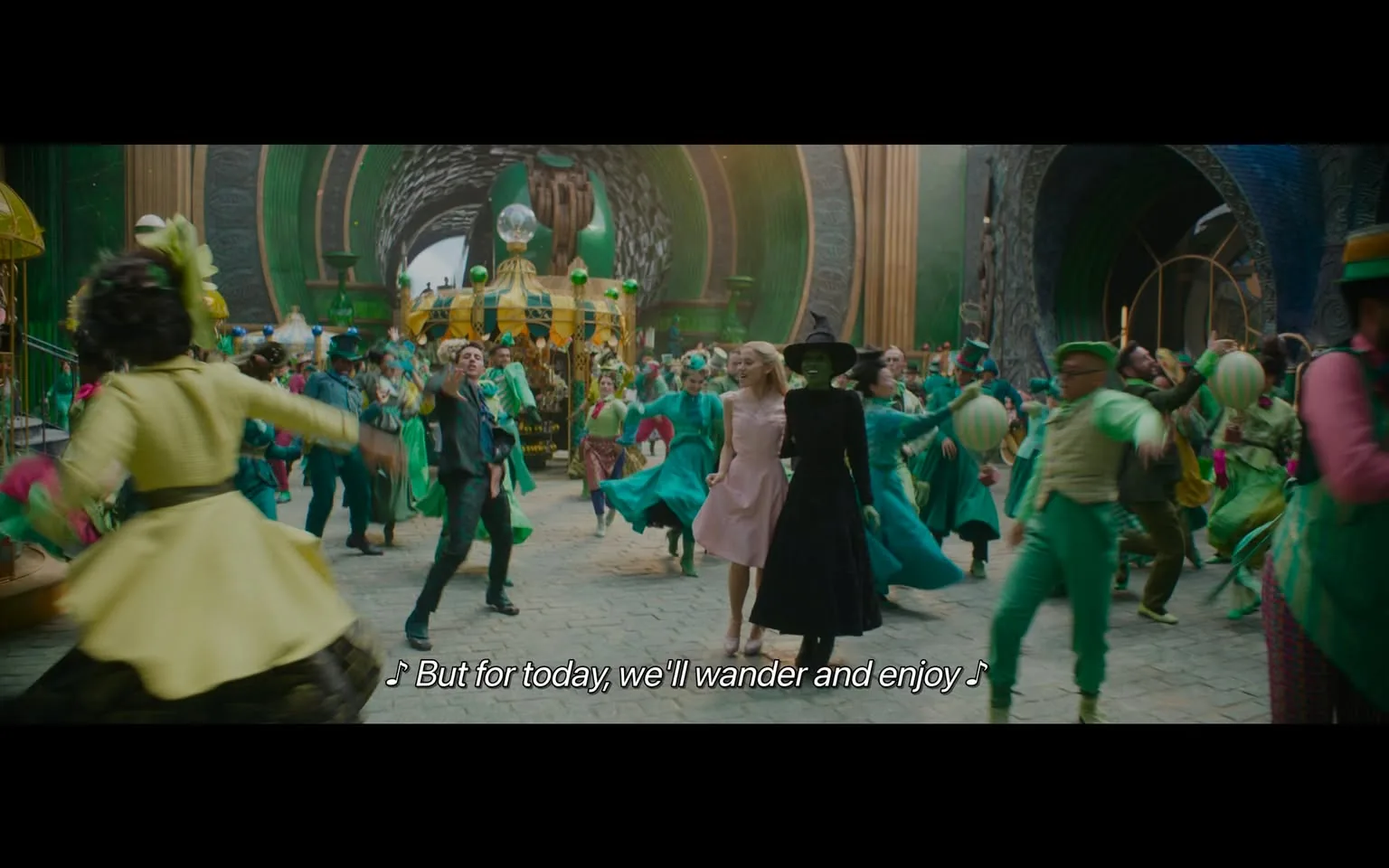 In a lively and colorful setting, a group of performers is engaged in a dance, creating a festive atmosphere. The scene features a mix of characters dressed in various shades of green and other bright colors, suggesting a whimsical theme. Two central figures stand out: one in a black dress and a pointed hat, and another in a light pink dress. They appear to be enjoying the moment, smiling and interacting with the surrounding dancers. The background is filled with elaborate decorations, including a carousel-like structure and green orbs, enhancing the magical ambiance. The lyrics visible at the bottom suggest a theme of enjoyment and wandering, fitting the joyful context of the performance.