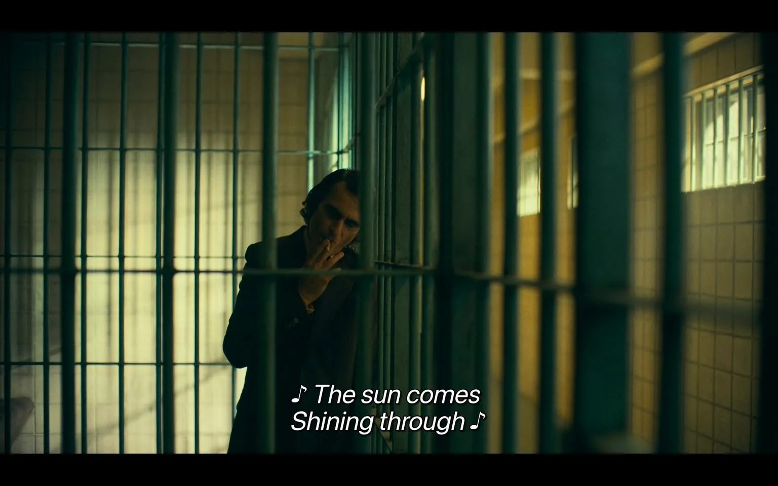 In a dimly lit prison cell, an unidentified adult stands near the bars, appearing contemplative as they smoke a cigarette. The individual is dressed in a dark suit, with their hair styled in loose waves. The setting is characterized by the stark, metallic bars of the cell, which create a sense of confinement. The walls are a muted color, and there are small windows in the background allowing some light to filter through, casting a soft glow. The visible text in the image includes lyrics that suggest a reflective mood: "The sun comes shining through." The overall atmosphere conveys a mix of melancholy and hope, typical of a narrative exploring themes of isolation and redemption.