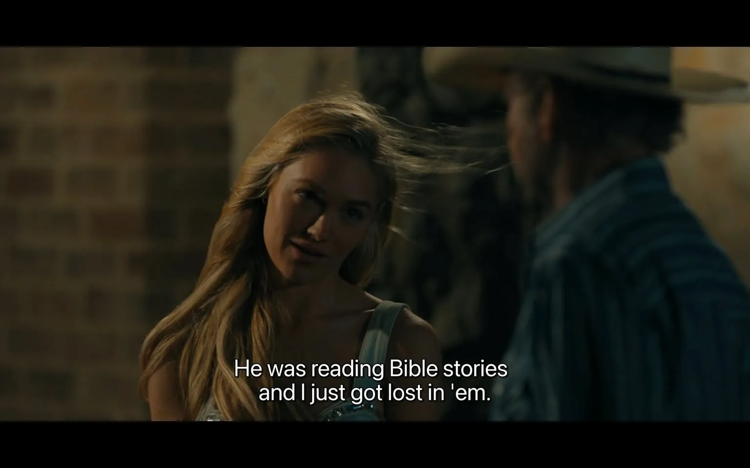 In the scene, a woman with long, flowing blonde hair is speaking to a man, who is partially visible and wearing a cowboy hat. The woman appears to be in her twenties or thirties and is dressed in a light-colored top. She has a thoughtful expression as she recounts a memory about someone reading Bible stories, indicating a sense of nostalgia or reflection. The background features a rustic brick wall, suggesting an indoor setting that may be casual or intimate. The lighting is warm, contributing to the overall mood of the conversation. The dialogue displayed on the screen reads, "He was reading Bible stories and I just got lost in 'em," emphasizing the emotional connection she feels to the stories.