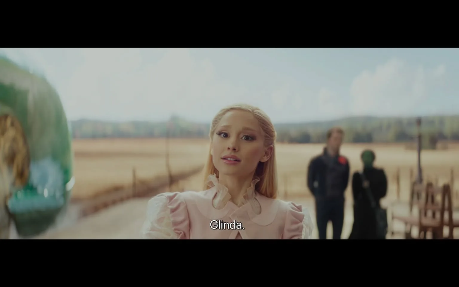 In the image, a character named Glinda is prominently featured, wearing a light pink dress with ruffled sleeves. She has long, straight blonde hair and is looking directly at the camera with a neutral expression. The background shows a vast outdoor landscape with fields and distant hills under a partly cloudy sky. In the distance, two other figures are visible, one in a dark outfit and another in a green outfit, but their details are less clear. The setting appears serene and rural, contributing to the overall whimsical atmosphere.