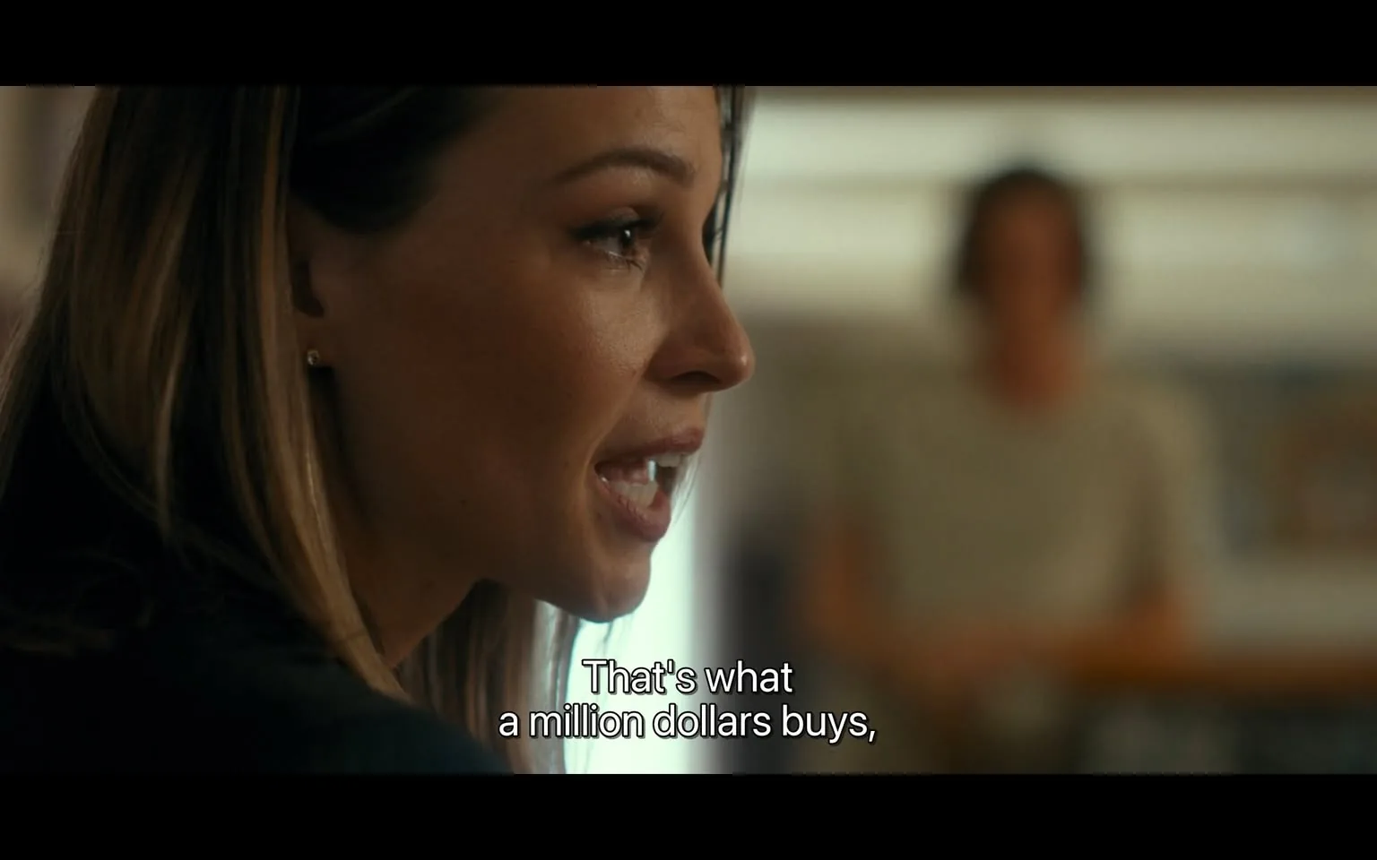 In the image, an unidentified adult woman is captured in profile, engaged in a conversation. She has long, dark hair and is wearing a black jacket. Her expression appears intense, suggesting she is making a significant point. The dialogue displayed on the screen reads, "That's what a million dollars buys," indicating a serious discussion about money or value. In the background, a blurred figure is present, but details about them are not discernible. The setting appears to be indoors, with soft lighting that creates a warm atmosphere.