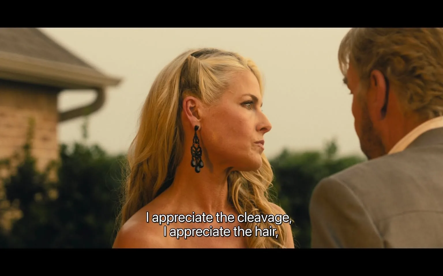 In the image, a woman with long, wavy blonde hair is seen in profile, wearing a strapless dress that reveals her shoulders and neckline. She has large, ornate black earrings that catch the light. Her expression appears serious as she engages in conversation with a man, whose face is partially visible. The background features a blurred outdoor setting, possibly a garden or patio, with greenery and a house structure. The lighting suggests it may be late afternoon or early evening. The text overlay indicates she is making a statement about appreciating certain physical attributes, which adds a layer of context to the interaction.